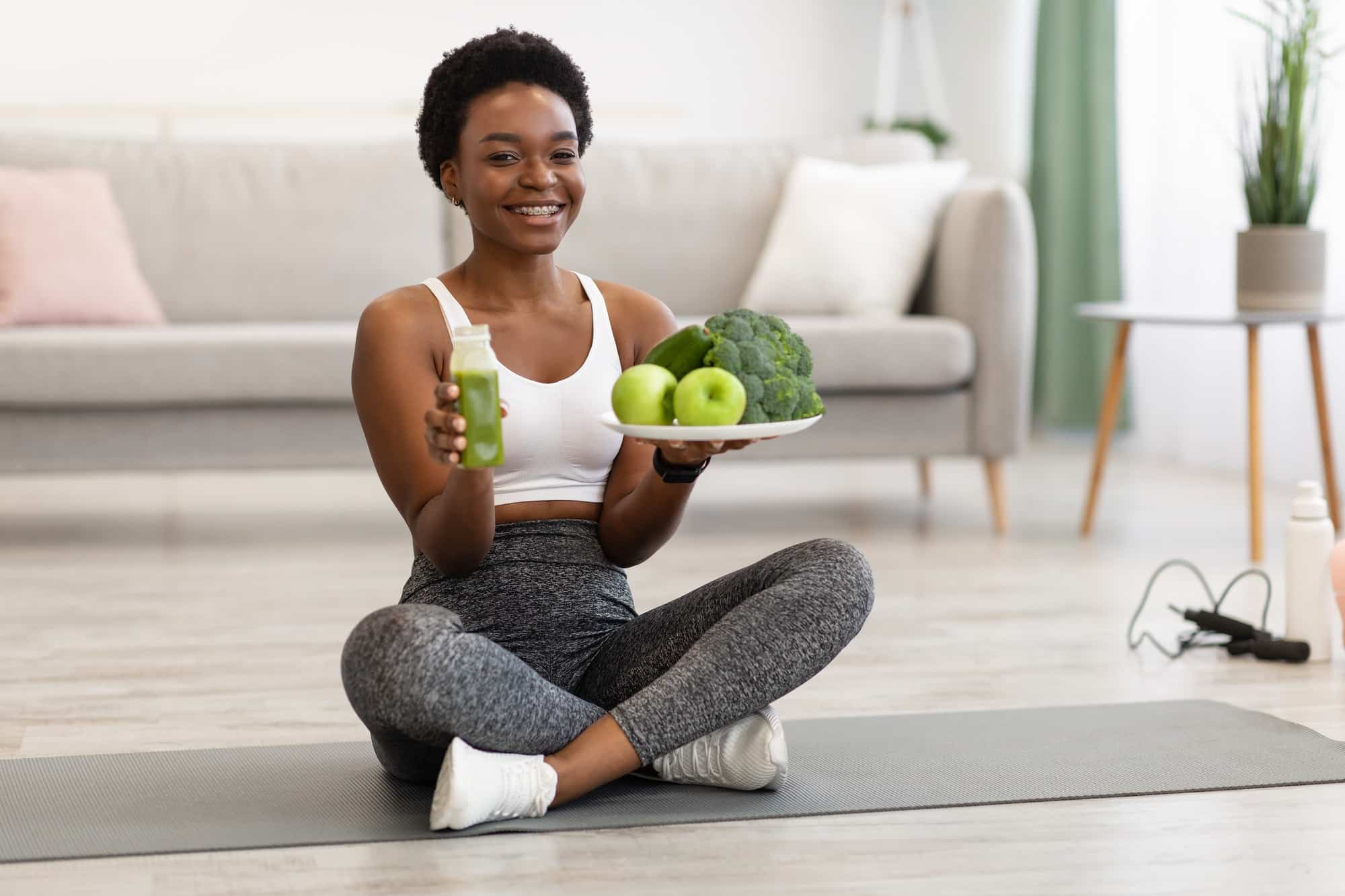 African Sporty Lady Holding Plate With Vegetables And Smoothie Indoors