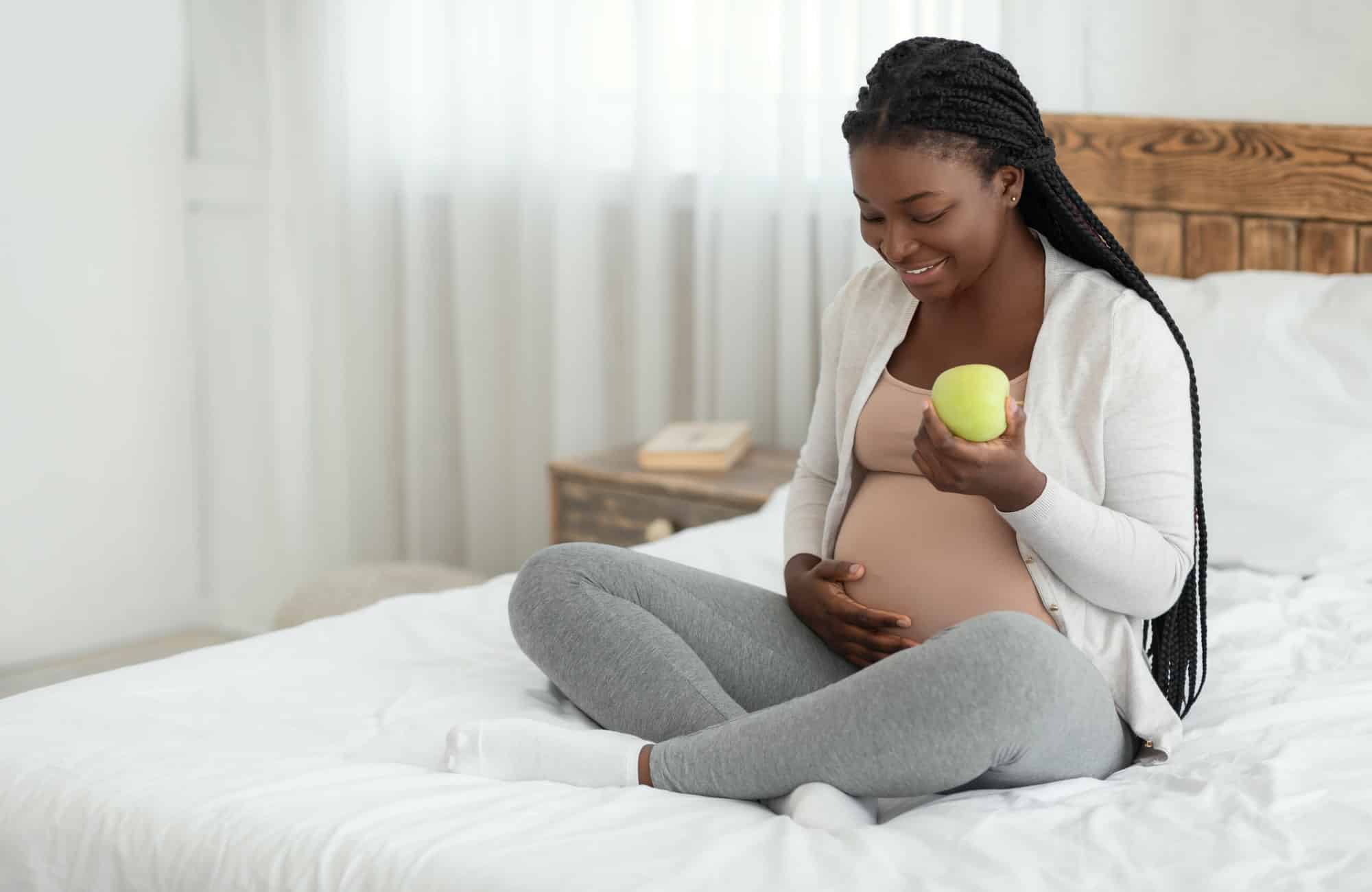Healthy Pregnancy Diet. Pregnant African Woman Eating Apple While Resting On Bed